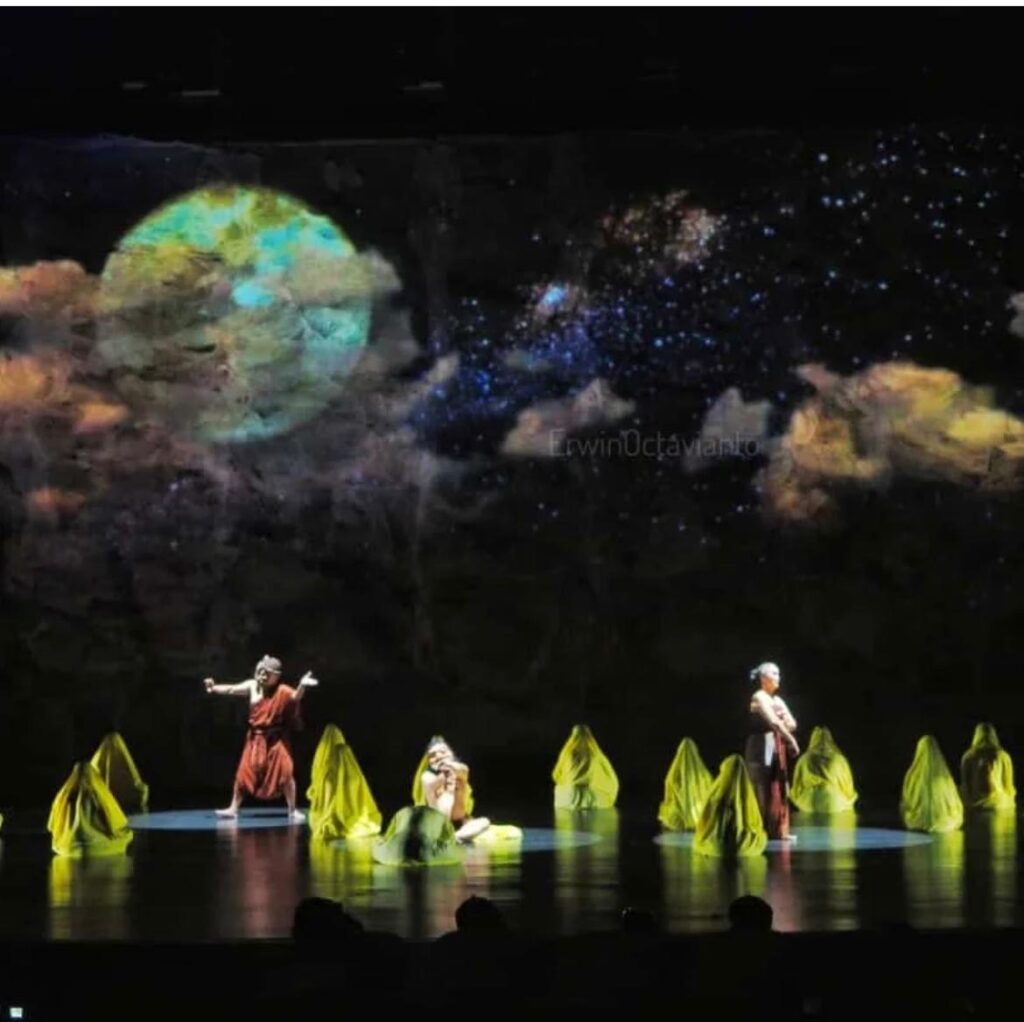 Konsep seni tari dan seni rupa catrawarta Stage performance with dancers in yellow cloaks surrounding two performers in brown set against a colorful space like projection of planets and stars behind them